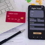 Office desk with credit card, smartphone with online airline ticket booking application on screen, laptop, paperwork, and pen